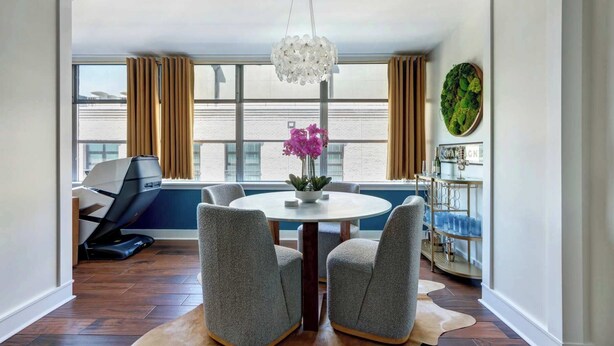A modern dining area with a round table, two grey chairs, a chandelier, a potted orchid, and a bar area with a green wall decoration.