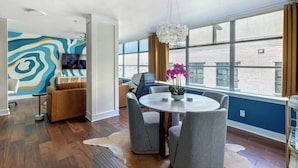 A modern living room with a round table, grey chairs, a large window with curtains, and a colorful abstract wall.
