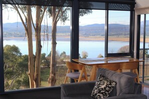 A room with a large window offering a view of a lake and distant mountains, a wooden table, and a grey sofa with a patterned pillow. - The Tamar Ridge Lookout 2Bed 2Bath Suite (Grindelwald)