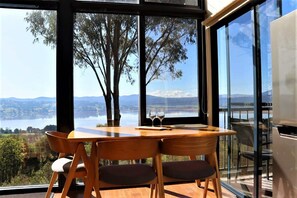A dining area with a table and chairs, large windows with a view of a lake and trees, and a modern interior design. - The Tamar Ridge Lookout 2Bed 2Bath Suite (Grindelwald)
