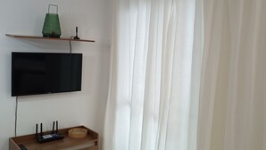 A room with a flat-screen TV mounted on the wall, a wooden console table with a remote and a router, and a white curtain covering the window.