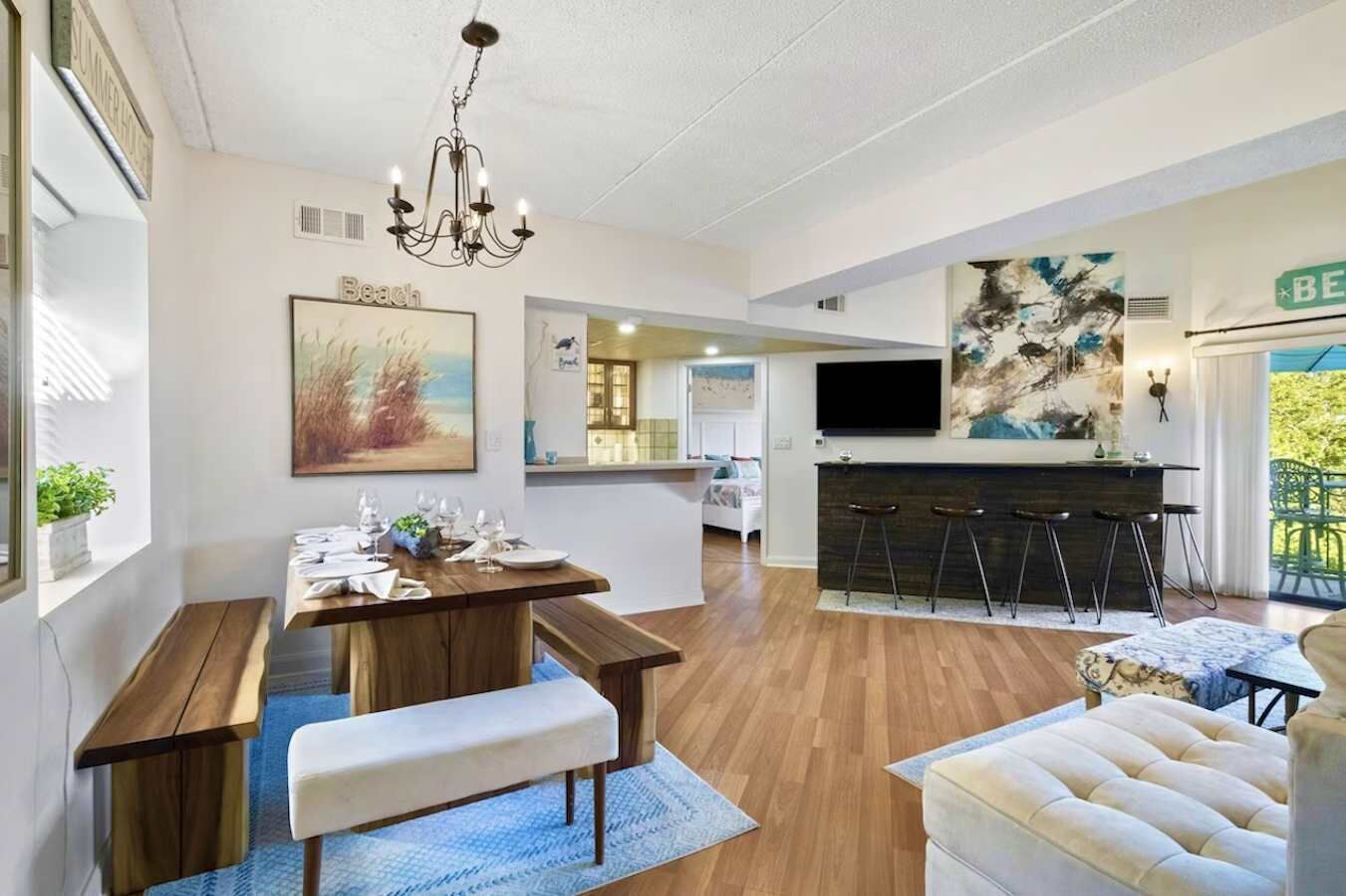 A modern living room with a dining area, a kitchen, and a bar area. There is a large painting on the wall, a chandelier, and a TV mounted above the bar.