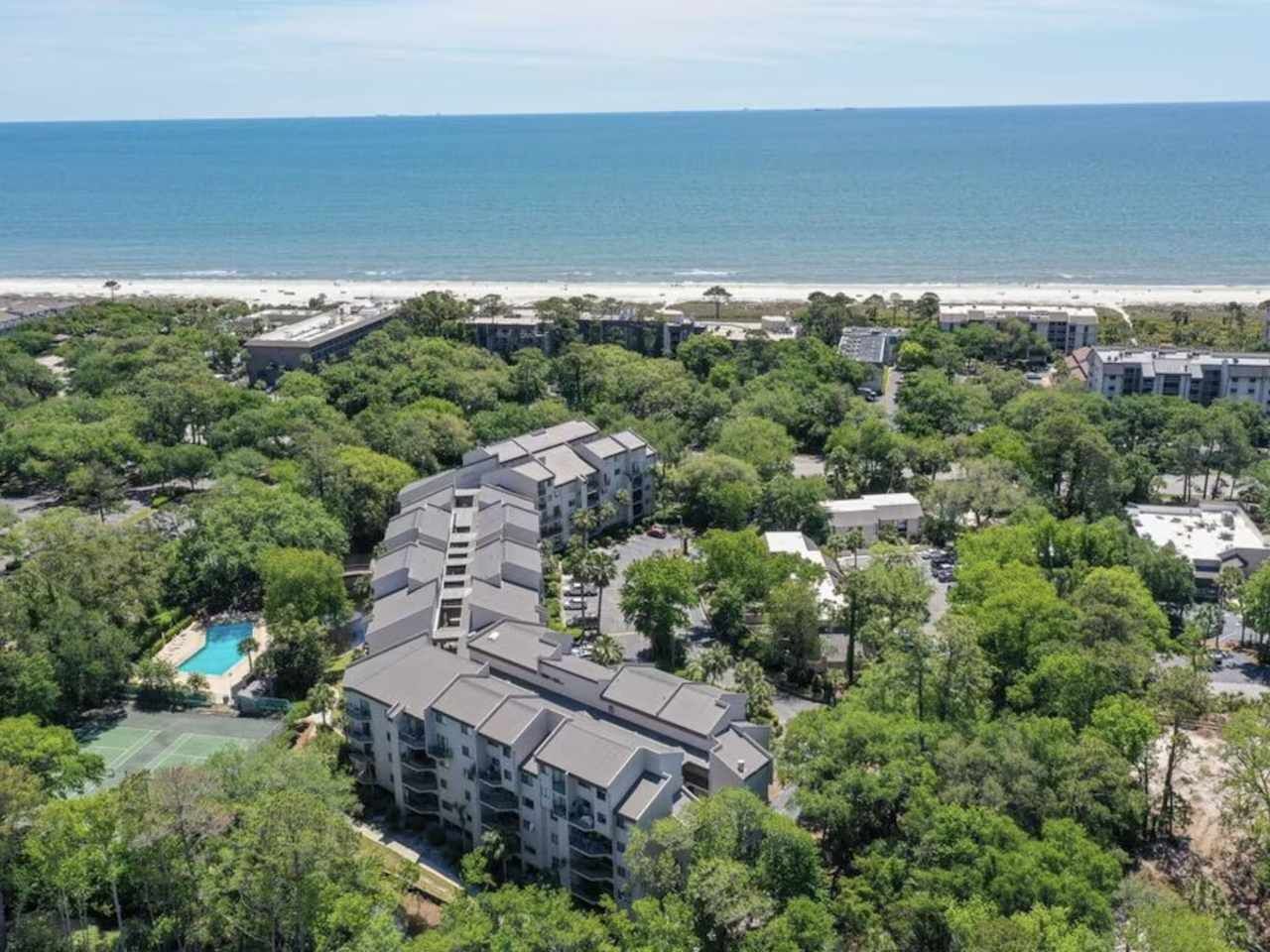 Un hôtel à plusieurs étages doté d’une piscine entourée d’arbres, situé près d’une plage.