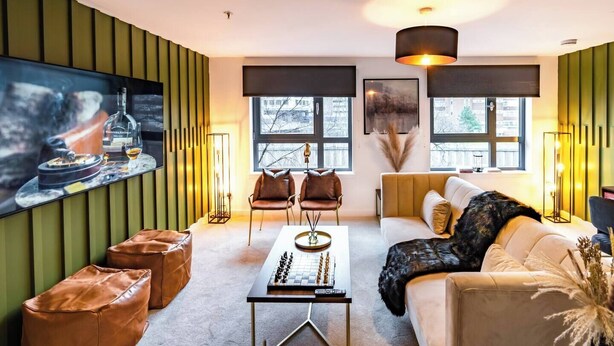 A modern living room with a sofa, two chairs, a coffee table, and a large painting. There are windows with views of the outside, a fireplace, and a floor lamp. - The Arran Suite - Your Stylish Scottish Getaway! (Glasgow)