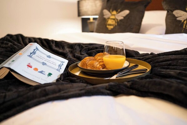 A bed with a black blanket, a book titled "I Never Knew That Scotland" by Christopher Williams, a plate with croissants and orange juice, and a lamp in the background. - The Arran Suite - Your Stylish Scottish Getaway! (Glasgow)