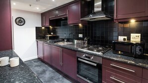 A modern kitchen with dark maroon cabinets, a black tiled backsplash, stainless steel appliances, and a built-in oven. - The Arran Suite - Your Stylish Scottish Getaway! (Glasgow)