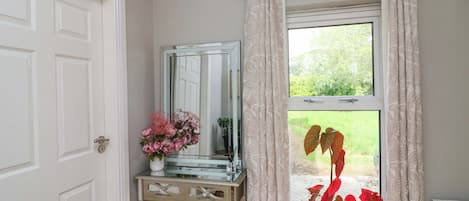 A room with a white door, a glass console table with a mirror, a potted plant, and a window with curtains.