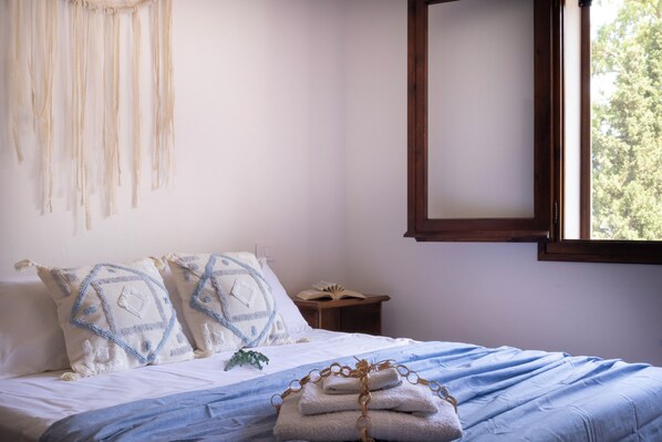 Una cama bien hecha con una colcha azul y blanca, dos almohadas decorativas, una mesita de noche con un libro y una ventana con vistas a árboles.
