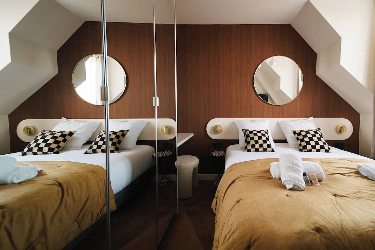 A hotel room with two beds, each with a checkered pillow, a round mirror, and a wooden headboard.