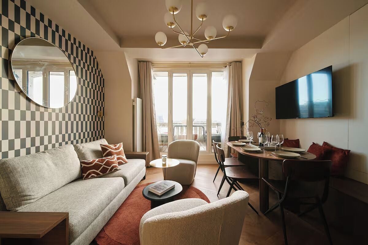 A modern living room with a checkered wall, a round mirror, a sofa, a coffee table, a dining area with chairs and a TV mounted on the wall.