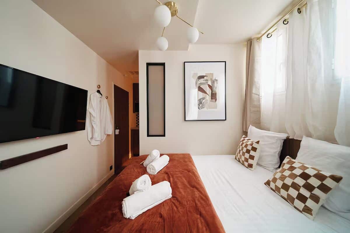 A hotel room with a large flat-screen TV, a bed with a brown headboard, white bedding, and checkered pillows.