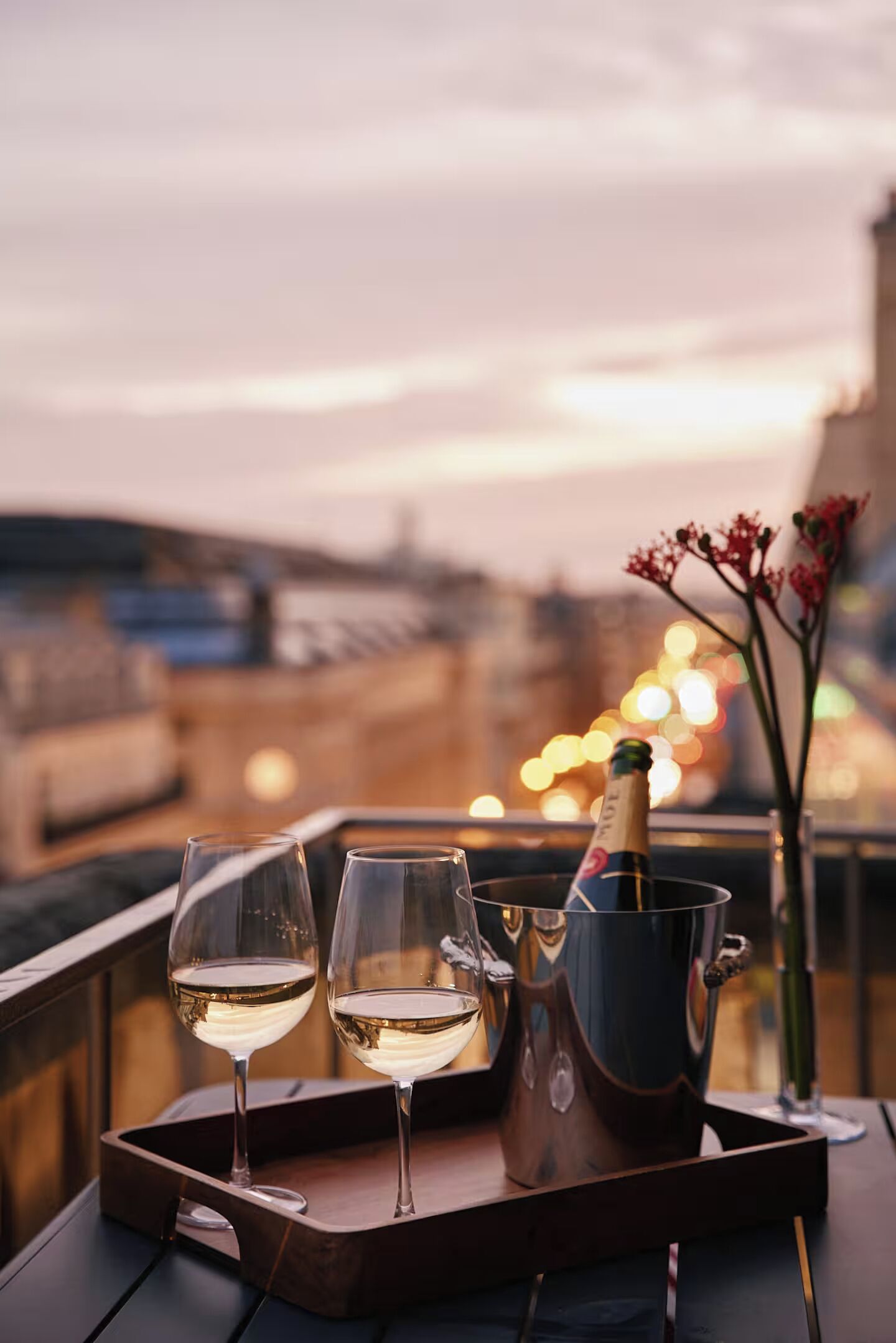 A tray with two glasses of white wine and a bottle of champagne on a balcony with a city view at dusk.