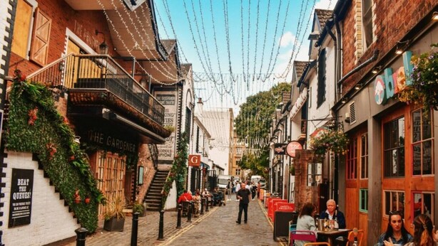 A cobblestone street lined with brick buildings, some with balconies and greenery. There are people walking and sitting at outdoor tables, suggesting a lively atmosphere.