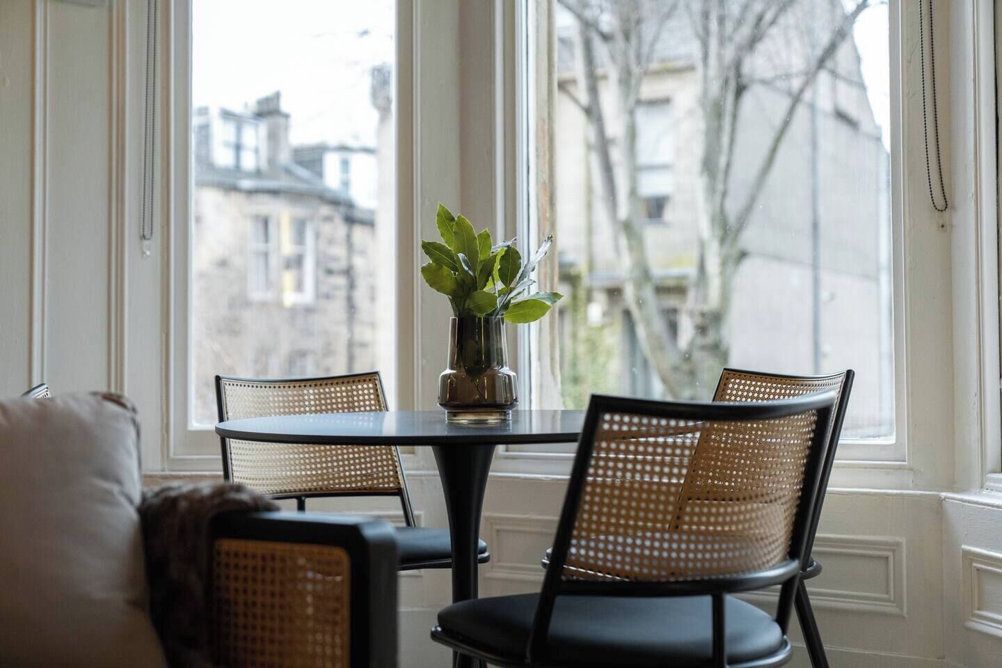 A dining area with a round table and four wicker chairs, a vase with green leaves, and a view of buildings through large windows.