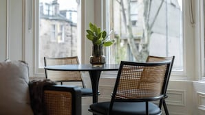 A dining area with a round table and four wicker chairs, a vase with green leaves, and a view of buildings through large windows.