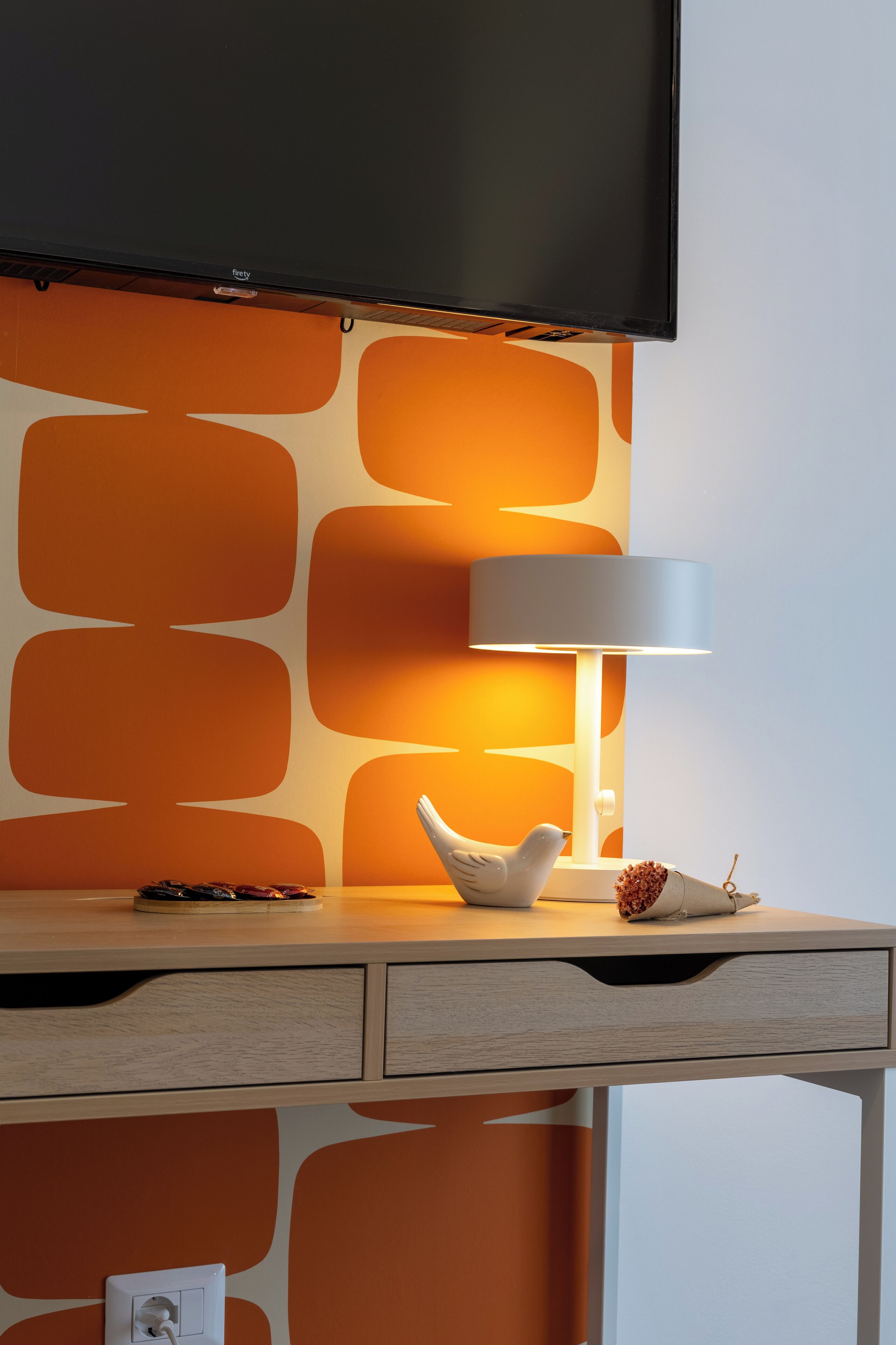 A modern room with a flat-screen TV mounted above a desk, a white lamp, and a decorative bird sculpture.