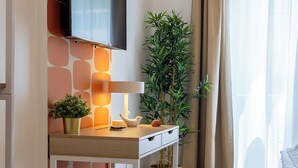 A modern living room with a flat-screen TV mounted on a wall, a wooden desk with a lamp, a potted plant, and a large window with curtains. - Cornelia Vatican Suites (Rome)
