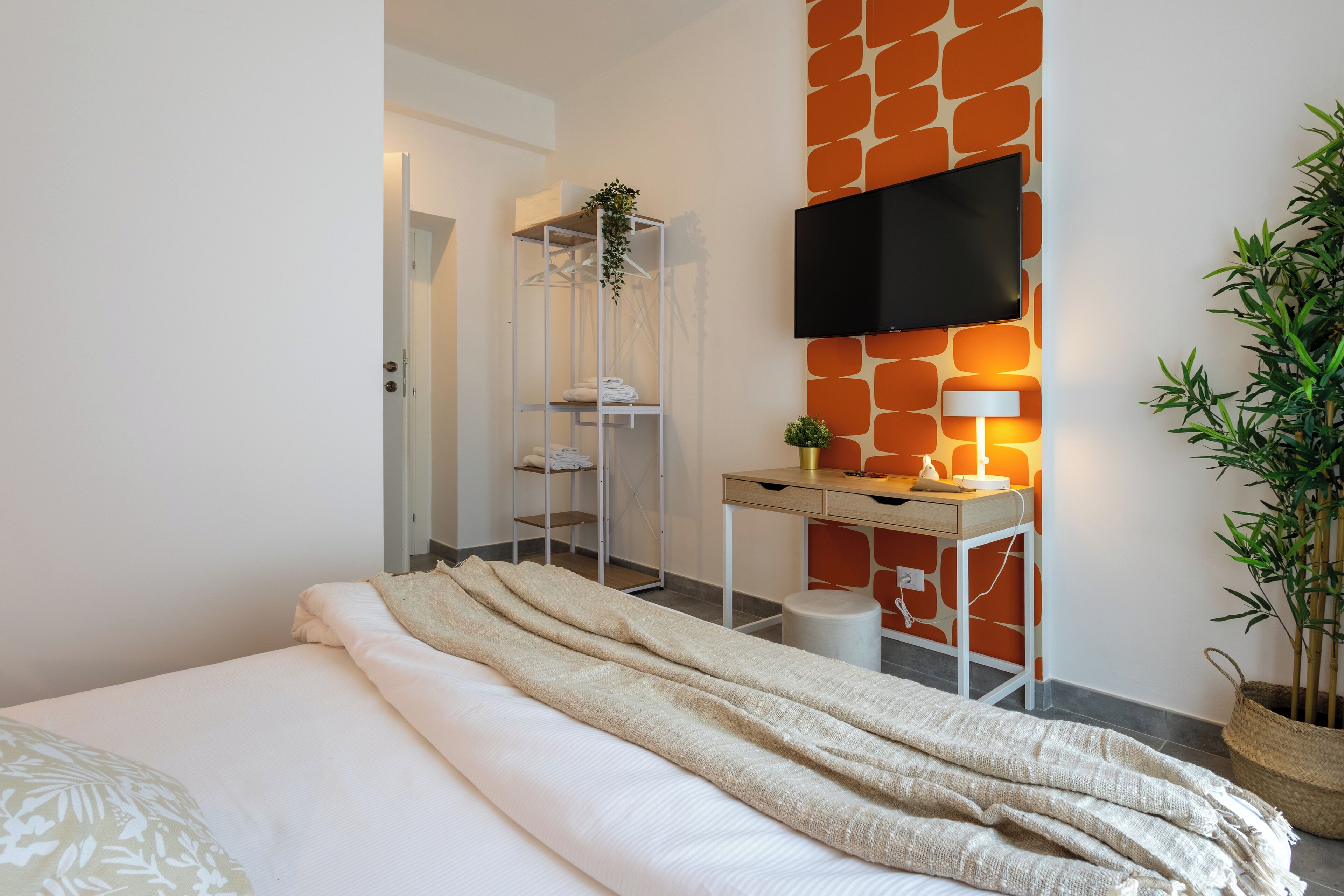 Ein modernes Hotelzimmer mit Bett, Schreibtisch mit Lampe, Fernseher und Pflanze.