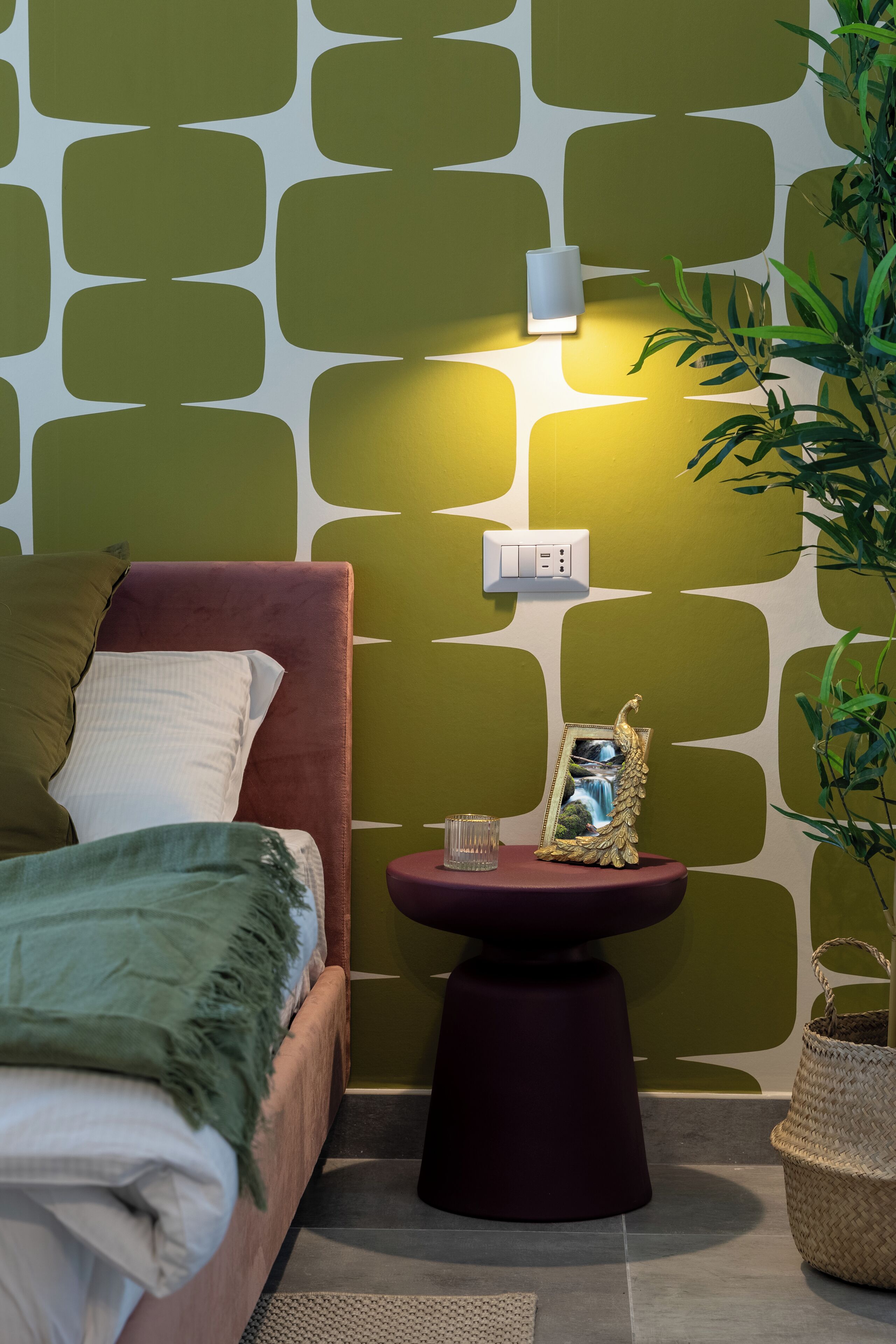 A bedroom with a bed, a bedside table with a picture frame, a wall-mounted light, and a potted plant.