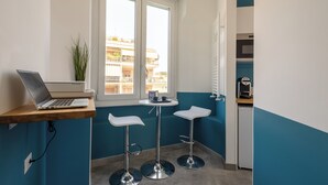 A small kitchenette with a laptop on a wooden shelf, two white bar stools, a small table, and a window overlooking a building. - Cornelia Vatican Suites (Rome)