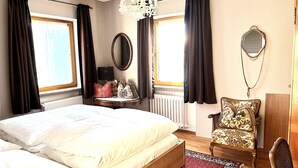 Een slaapkamer met een groot bed, een nachtkastje met een lamp, een stoel, een spiegel en een raam met gordijnen.