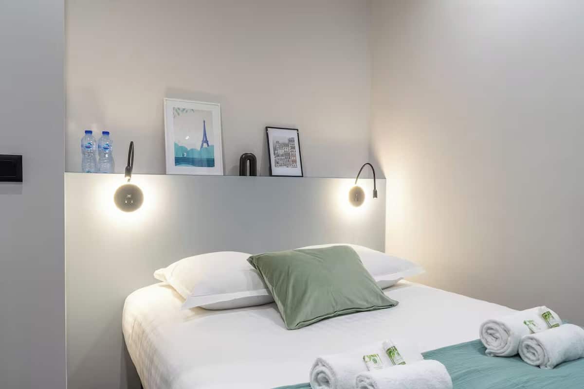 Un lit bien fait, agrémenté d’oreillers blancs et verts, d’une couvre-lit turquoise et de serviettes enroulées posées sur le côté. Deux tableaux encadrés sont accrochés au-dessus du lit, et on aperçoit un luminaire fixé au mur.