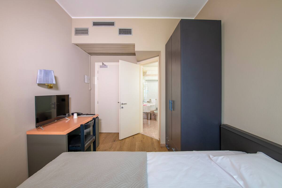 A hotel room with a bed, a desk with a chair, a television, and a bathroom visible through an open door.