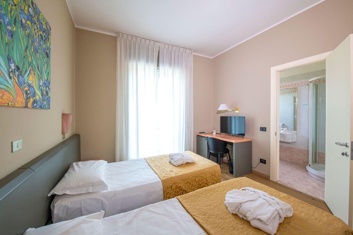 A hotel room with two beds, a desk with a TV, a painting on the wall, and a bathroom visible through an open door.