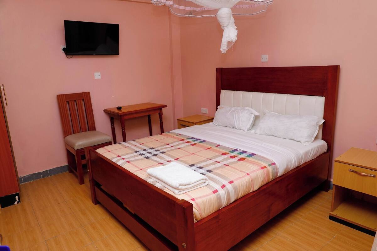 Ein Schlafzimmer mit einem hölzernen Bett, einem Nachttisch, einem Sessel, einem an der Wand montierten Fernseher und einem Moskitonetz über dem Bett.