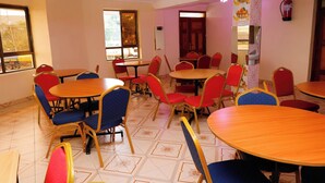 Un área de comedor con varias mesas redondas y sillas, un extintor en la pared y una ventana con vista a un edificio.