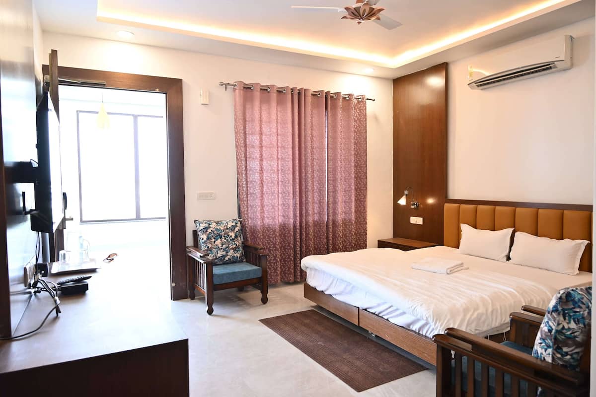 A hotel room with a bed, a desk with a computer, a chair, a window with curtains, and an air conditioning unit.