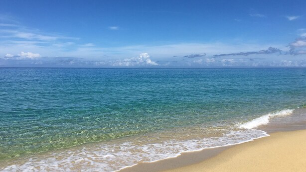 Une plage à l’eau claire, avec un rivage sablonneux et des nuages qui se dessinent à l’horizon.