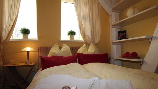 A hotel room with a bed, a bedside table with a lamp, two windows with curtains, a shelf with decorative items, and a small potted plant on the windowsill. - Hof Eckhorst, Apartment Sonnenschein (Güby)
