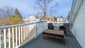 Une terrasse dotée de meubles en osier et d’une balustrade blanche, qui offre une vue sur un quartier résidentiel avec ses maisons et ses arbres.