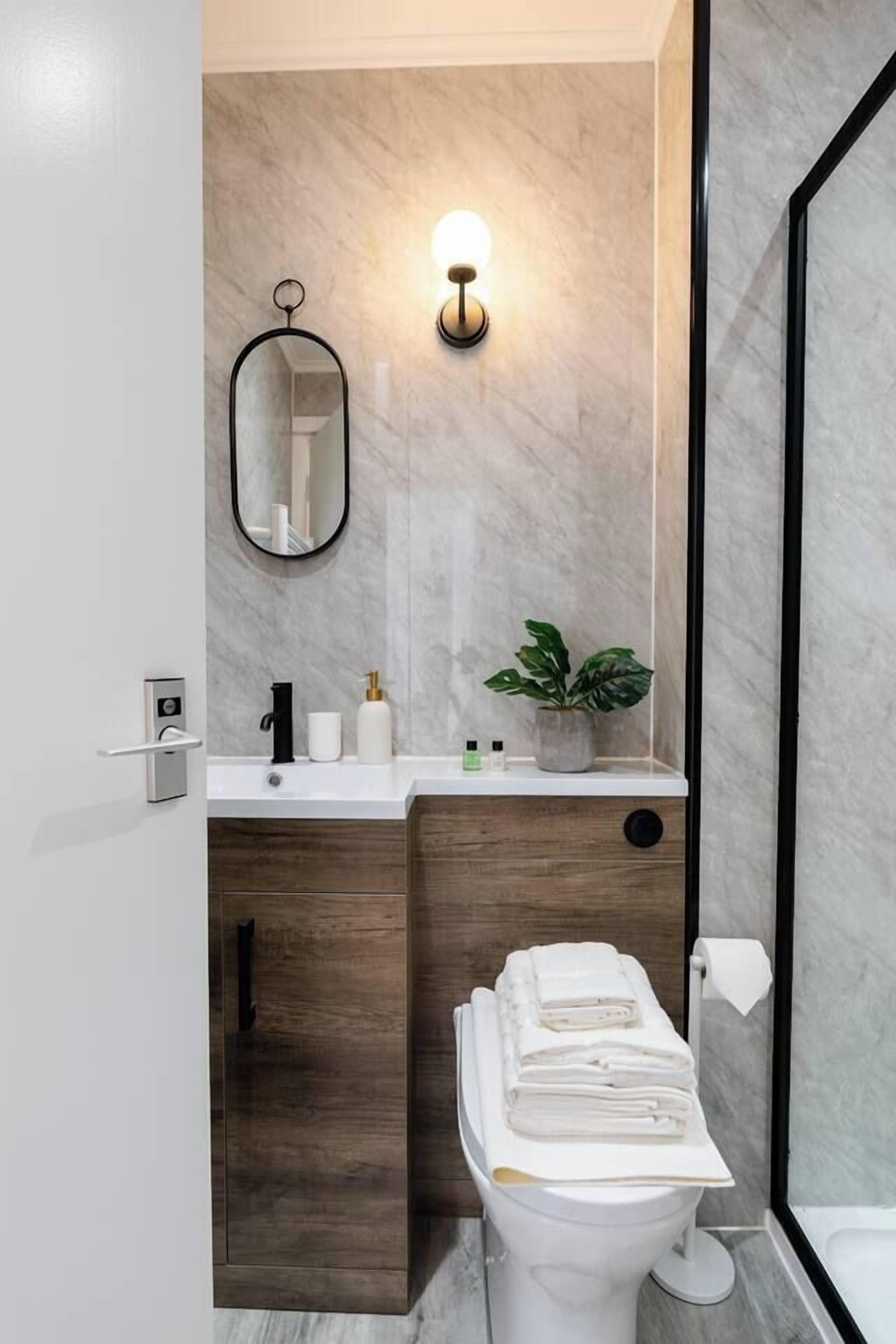 A modern bathroom with a marble wall, a wooden cabinet, a toilet with a towel stack, a wall-mounted mirror, and a light fixture.
