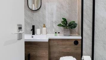 A modern bathroom with a marble wall, a wooden cabinet, a toilet with a towel stack, a wall-mounted mirror, and a light fixture.