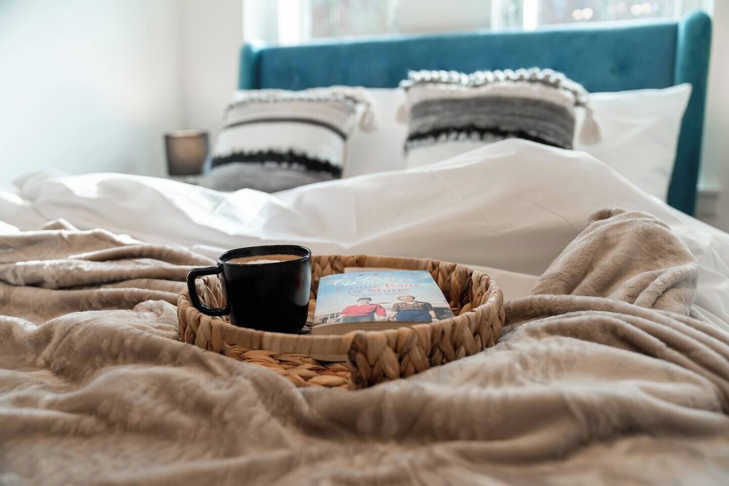 A bed with a coffee cup and a book on a woven tray.