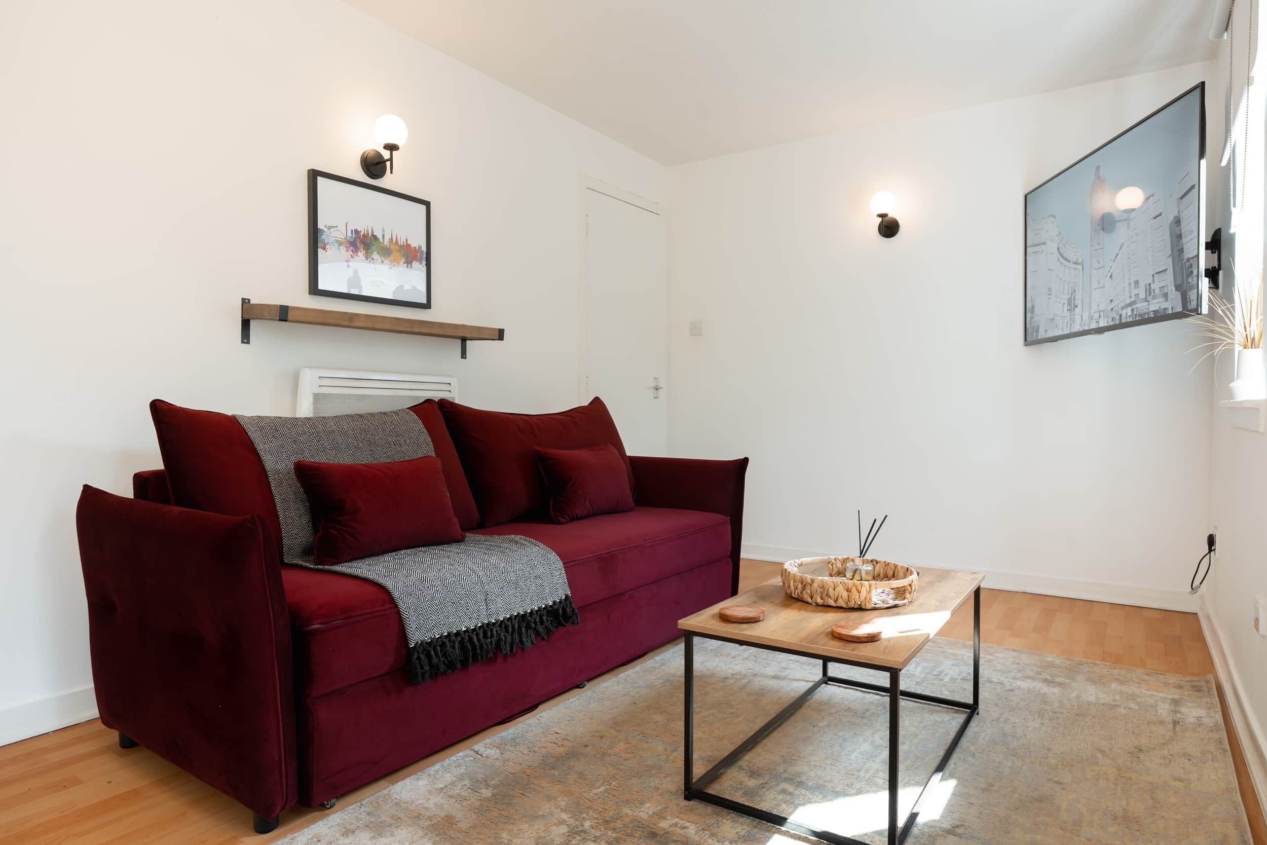 A red velvet sofa with a gray throw blanket, a wooden coffee table with a woven basket, and a framed cityscape picture on the wall.