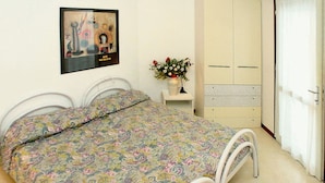 A bedroom with a bed, a wardrobe, a small table with a vase, and a picture on the wall. - Cozy Apartment Near the Beach With Parking Space Included (Lignano Sabbiadoro)