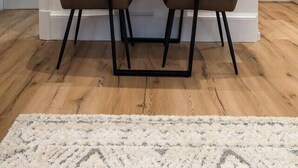 Un comedor con una mesa de madera, dos sillas, un jarrón con plantas secas y un cuadro en la pared.