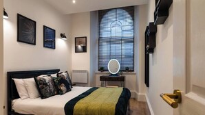 Un dormitorio con una cama, una mesita de noche con espejo, una ventana con persianas y una obra de arte colgada en la pared.