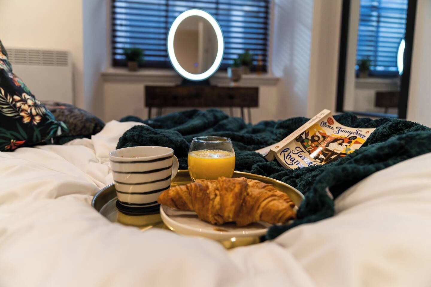 Un petit-déjeuner soigneusement préparé, servi sur un plateau posé sur le lit, avec une tasse à rayures, un verre de jus d’orange, un croissant et un magazine.