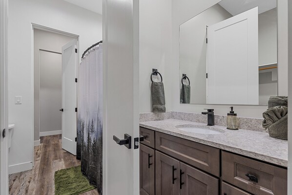 A bathroom with a white vanity, dark wooden cabinets, a large mirror, and a shower curtain with a forest design.