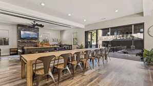 Een ruime eethoek met een lange houten tafel en moderne stoelen, een metselen haard en een groot raam met uitzicht op een balkon.