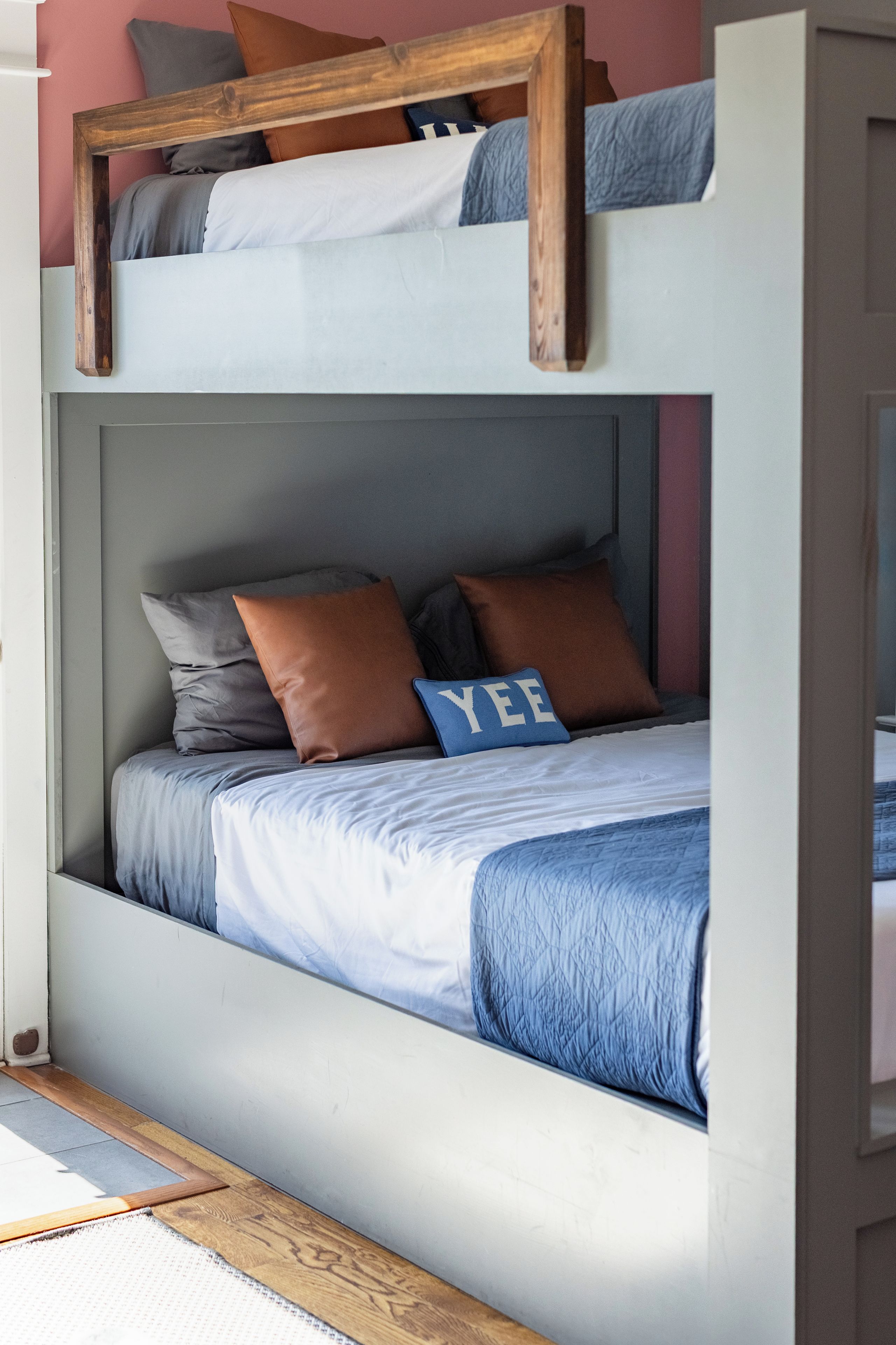 A bunk bed with a wooden frame, grey bedding, and a blue pillow with the word "YEE".