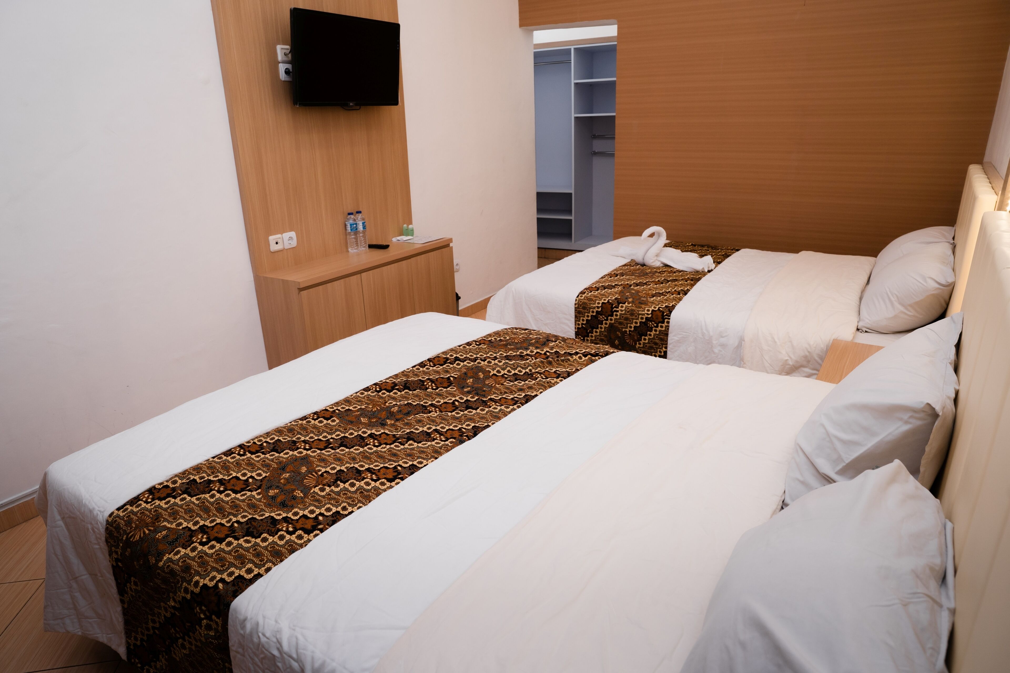 Une chambre d’hôtel avec deux lits, un téléviseur fixé au mur, une table de chevet en bois avec une bouteille et un verre, et une armoire contenant des vêtements suspendus.