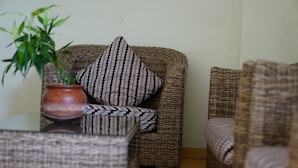 A wicker chair with a patterned pillow, a glass table with a potted plant, and a wicker sofa. - Grha Somaya (Yogyakarta)