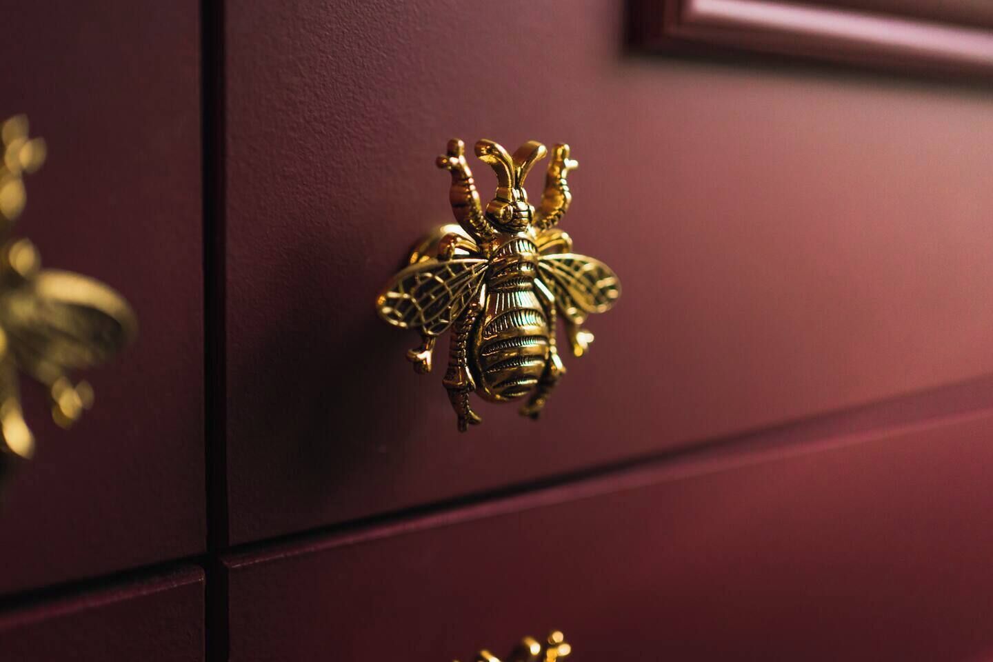 Ein Detailaufnahme eines goldfarbenen, bienenörmigen Griffs an einer dunkelroten Schublade.