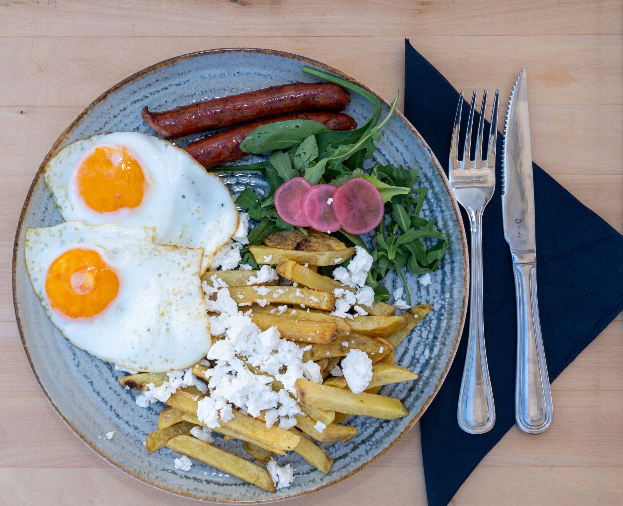 A plate with eggs, fries, and meat, accompanied by a fork and knife on a napkin.
