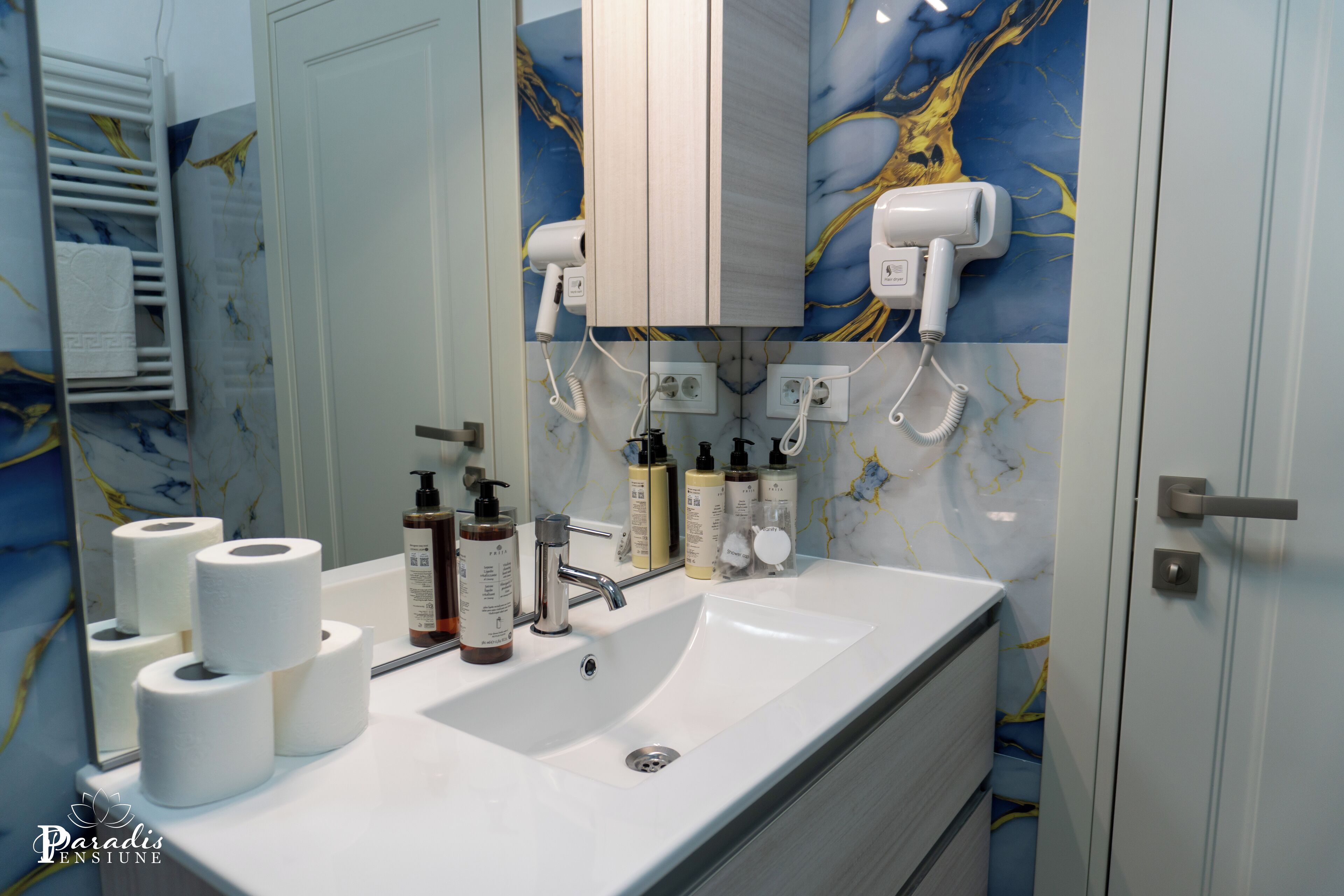 Un bagno con lavandino bianco, numerosi articoli da toilette, un asciugacapelli e un porta asciugamani.